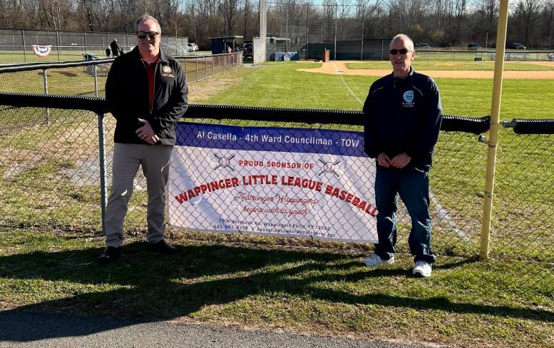 Two people holding a banner on a grassy field at a little league baseball complex.