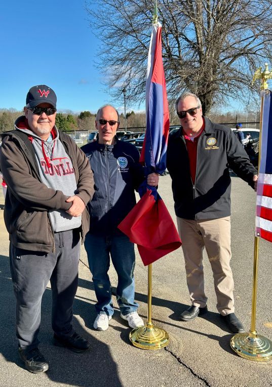 Three men posing outdoors with a flag on a sunny day