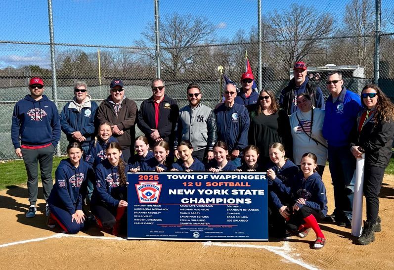 Baseball team posing with championship banner on a sunny field, wearing matching jackets and uniforms