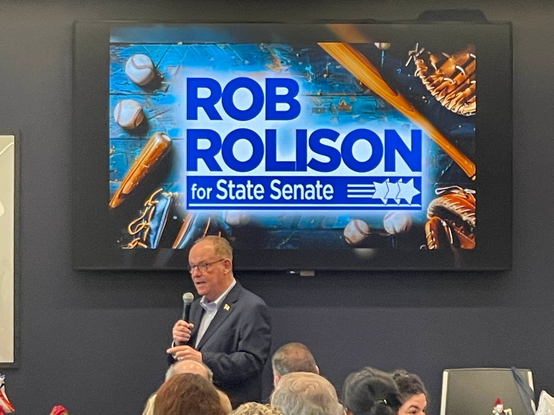 Man speaking on stage before a Rob Rolison for State Senate campaign sign