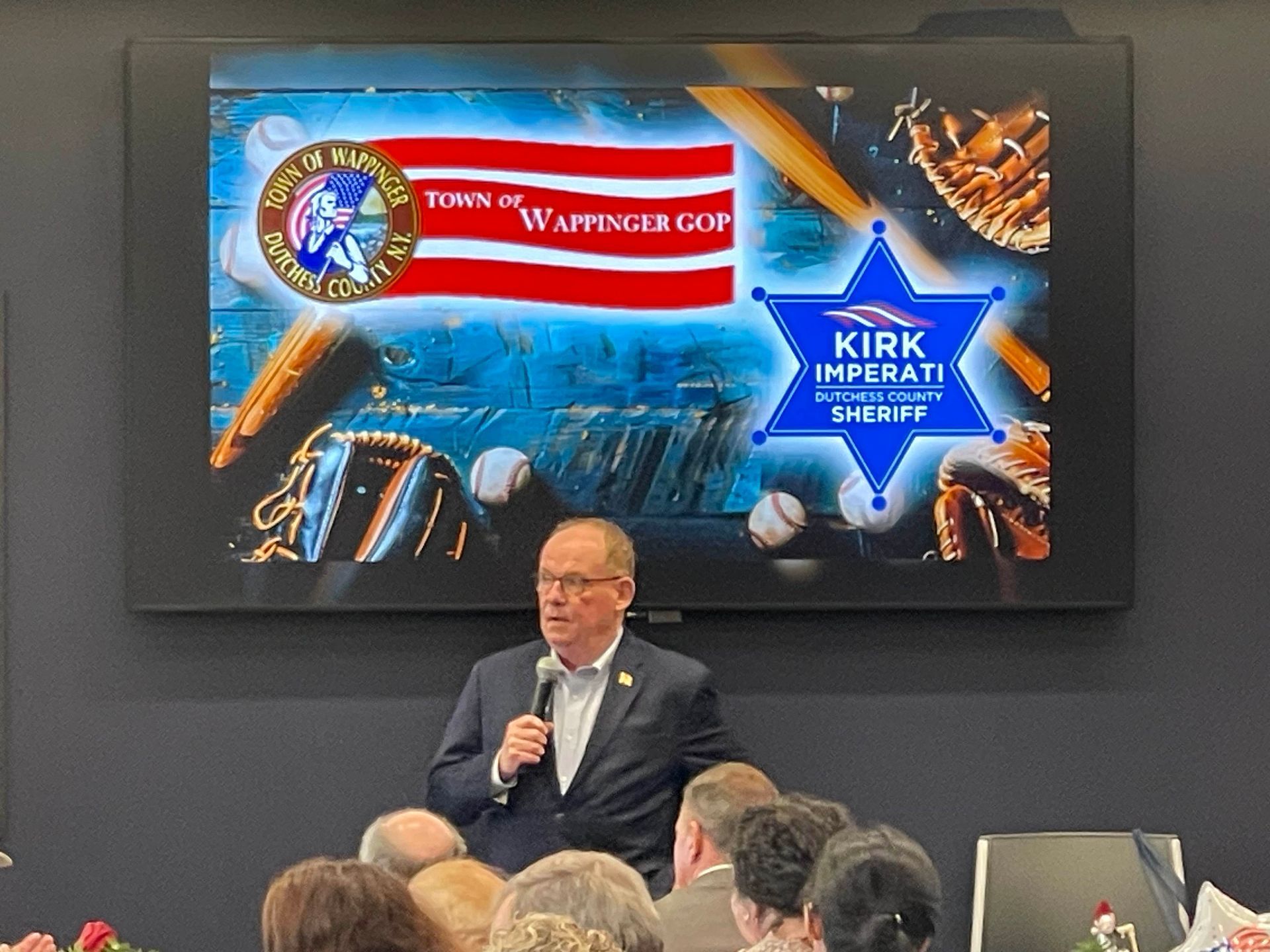 Man speaking at a podium before a large screen showing a graphic with a police badge and “Kirk” text