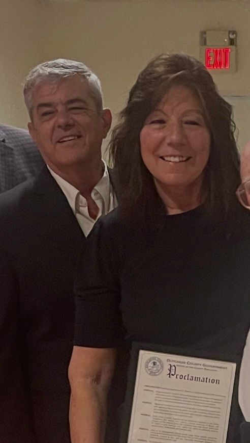 Two people smiling indoors, holding a framed certificate, with a red EXIT sign in the background.