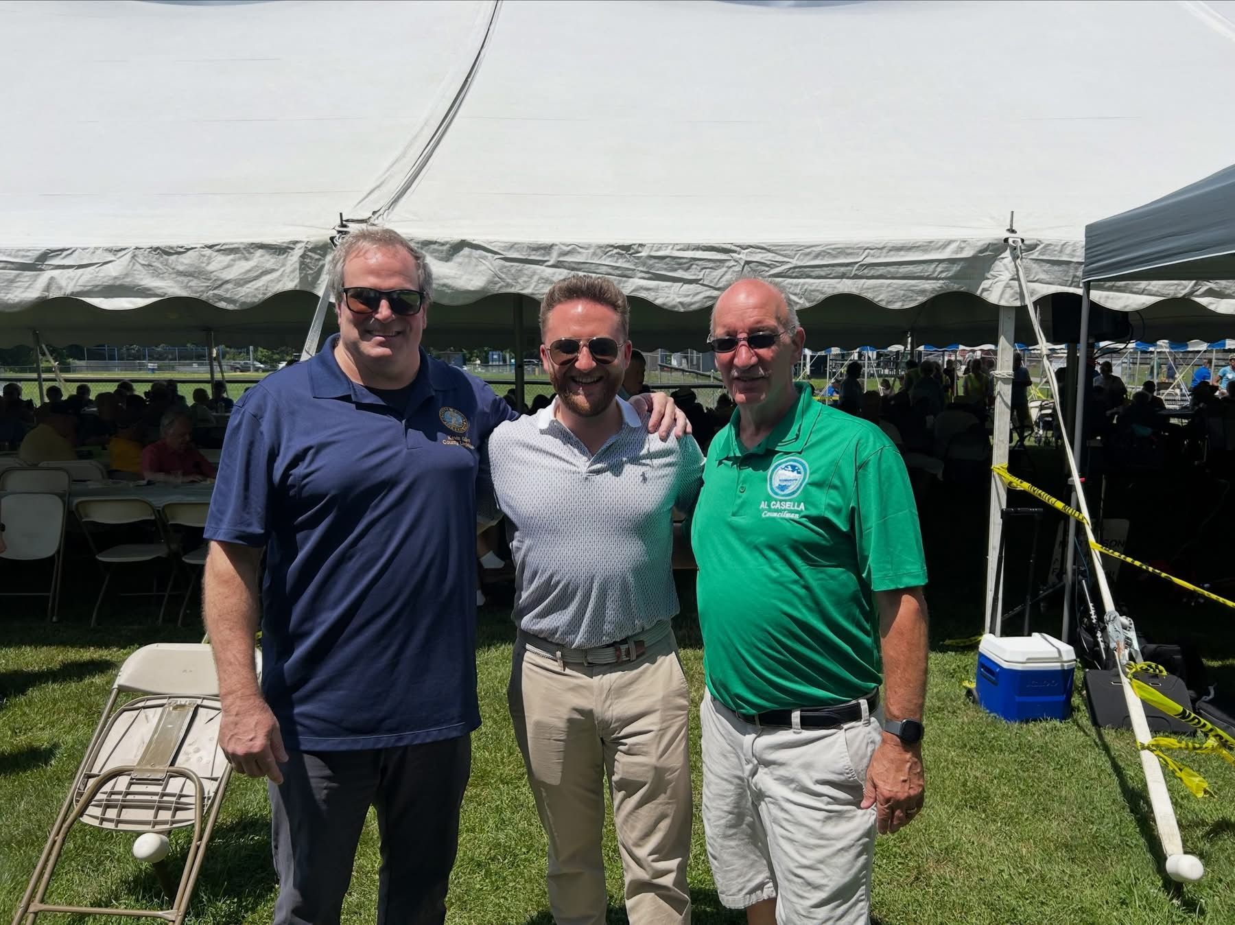 Three men standing together under a white event tent on a sunny lawn, smiling at the camera.