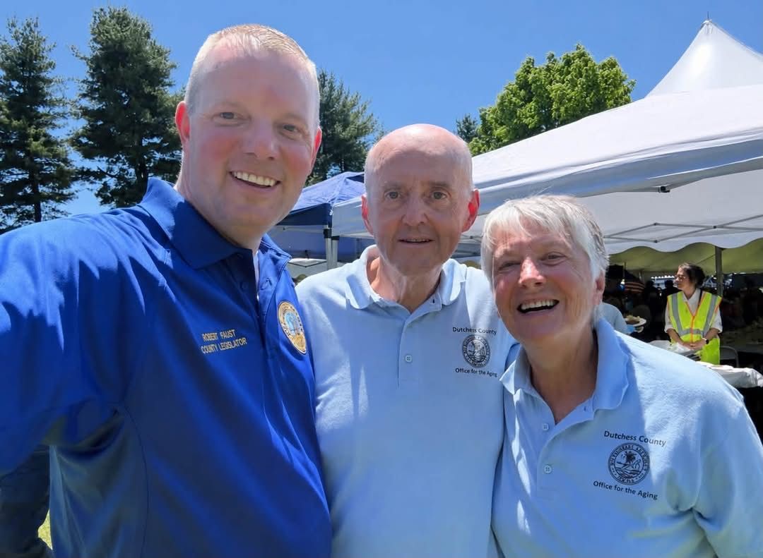 Three smiling people in blue shirts pose outdoors under a white canopy at an event.