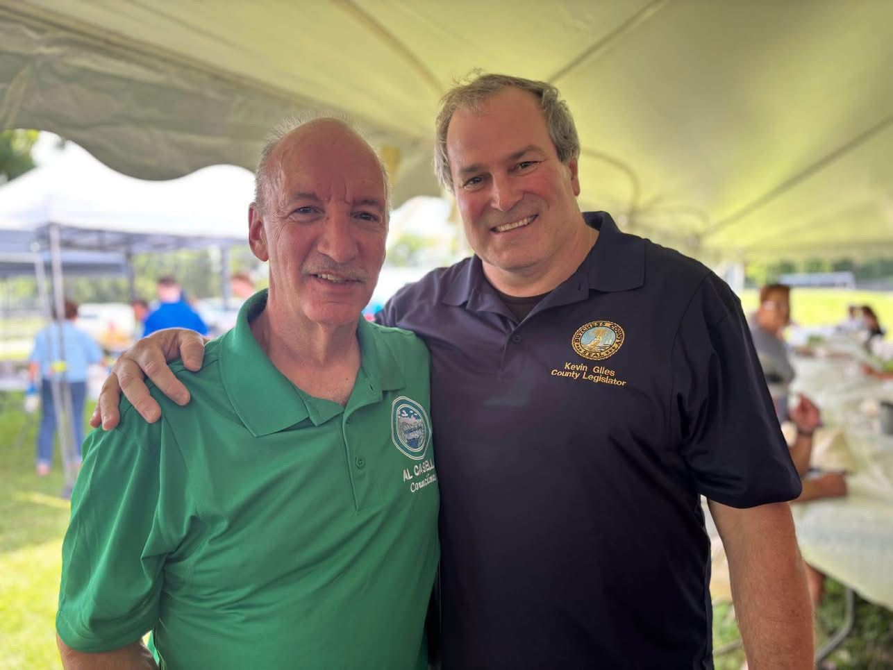 Two men smiling under a tent, one in a green shirt and one in a black polo, with an outdoor event in the background
