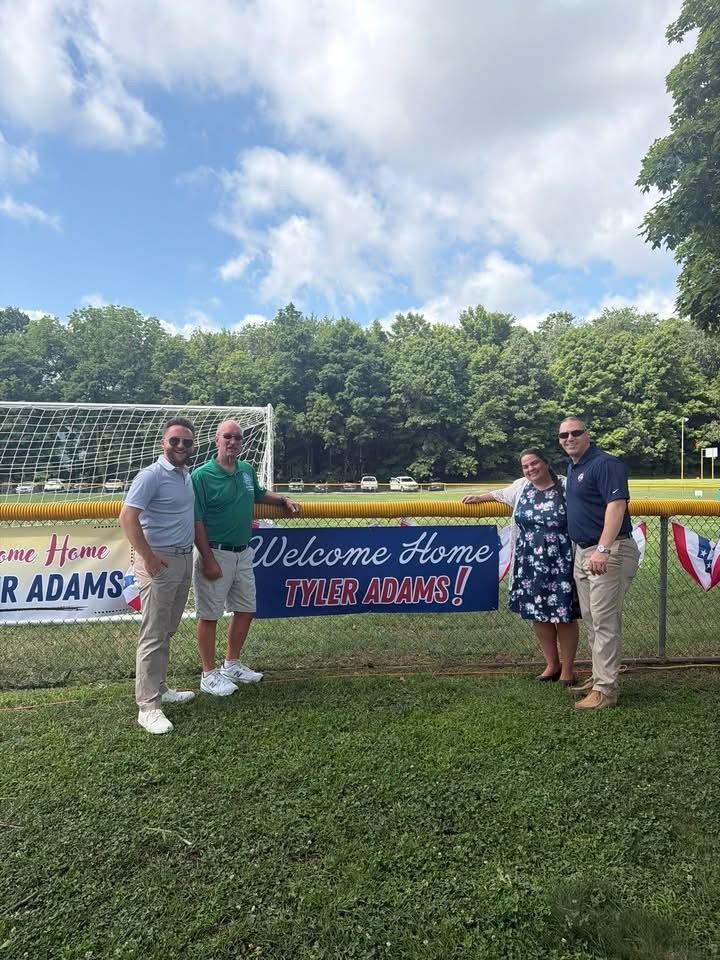 Three people posing by a “Welcome Home SSGR ADAMS” banner on a grassy field.