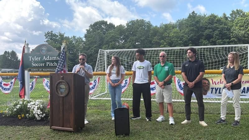 People speaking at a podium on a soccer field, with several others standing beside a goal and banners behind them.