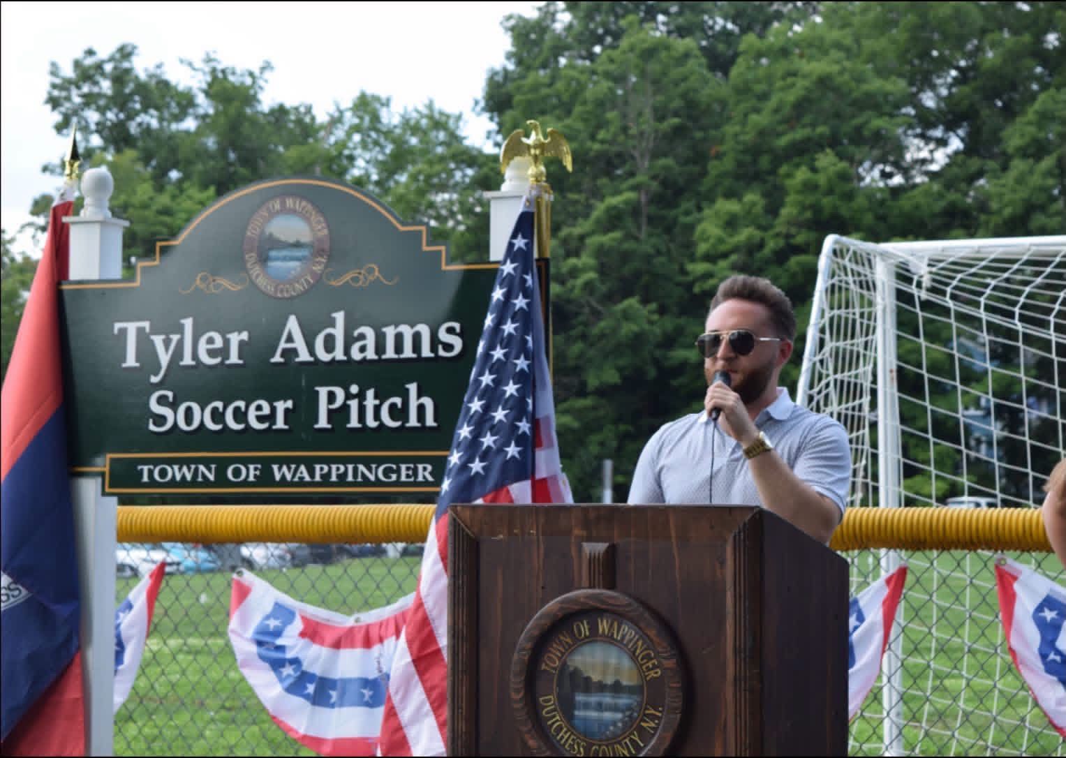 Speaker at podium beside Tyler Adams Soccer Pitch sign with U.S. flags and soccer goal backdrop