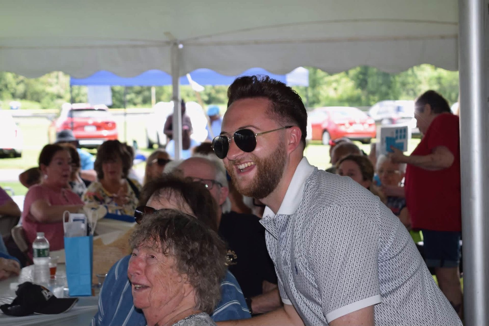 Smiling man in sunglasses leaning over a seated crowd under a white event tent