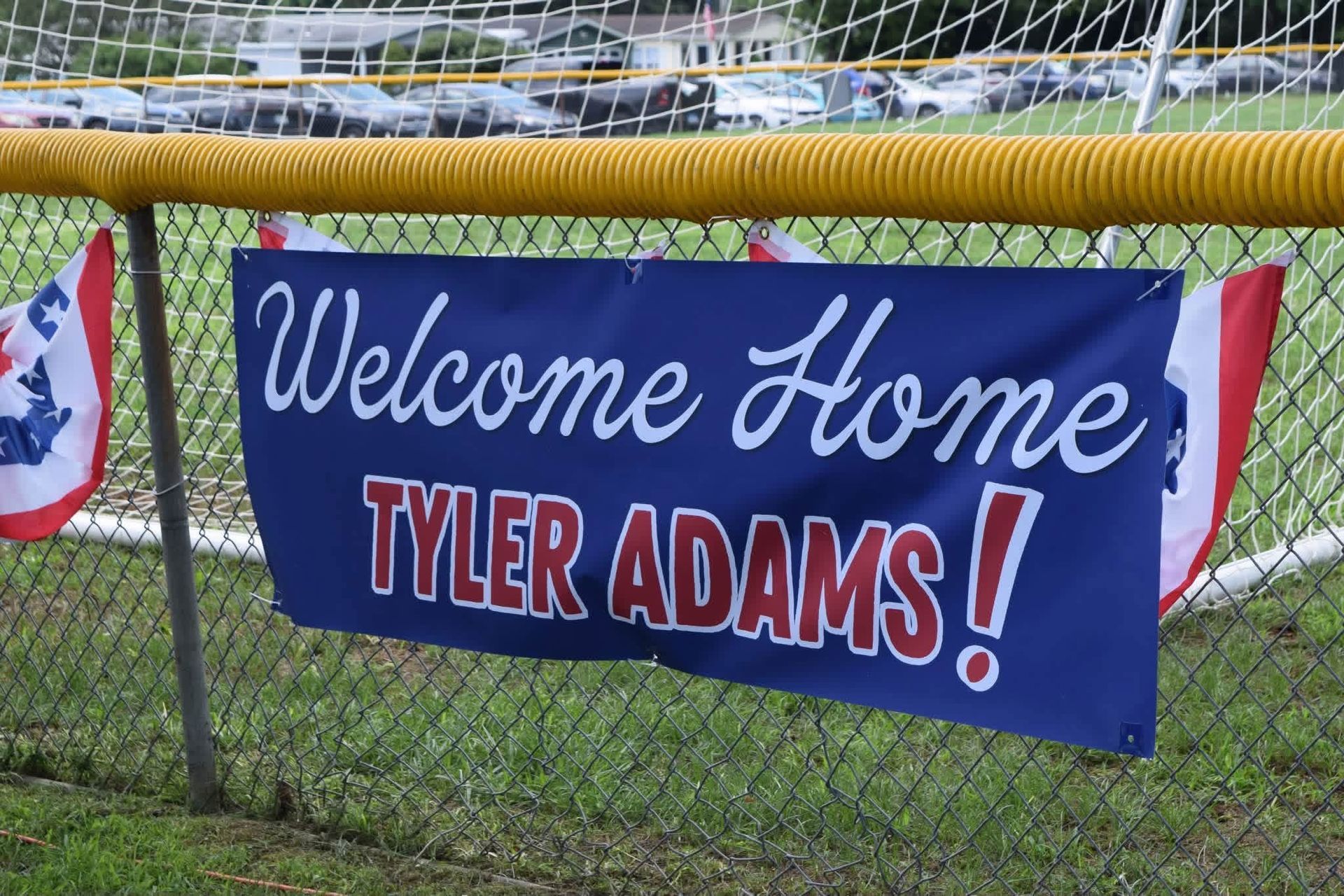 Blue Welcome Home Tyler Adams! banner hanging on a fence at a sports field