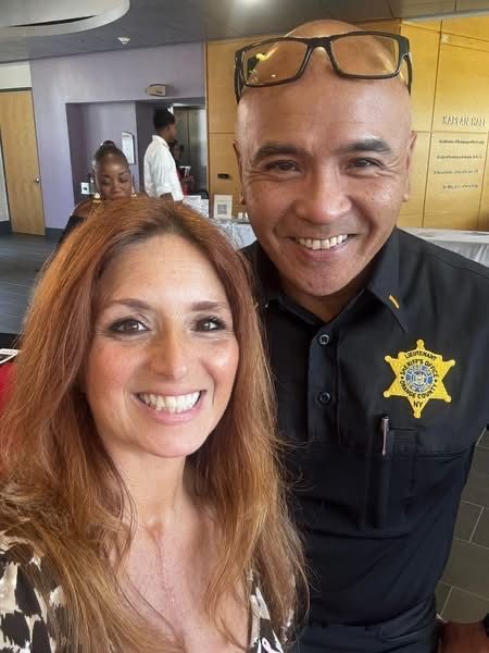 Two smiling people posing indoors, one in a black sheriff shirt with a gold badge.