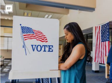 Person voting at a ballot booth with a U.S. flag and a sign