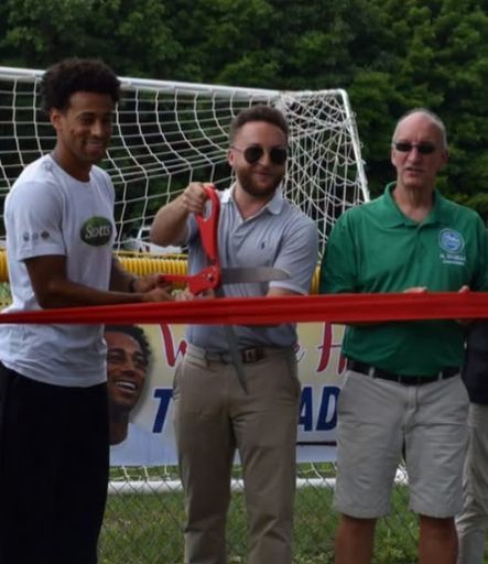 Three men at a ribbon-cutting ceremony on a soccer field, with a goal net in the background.