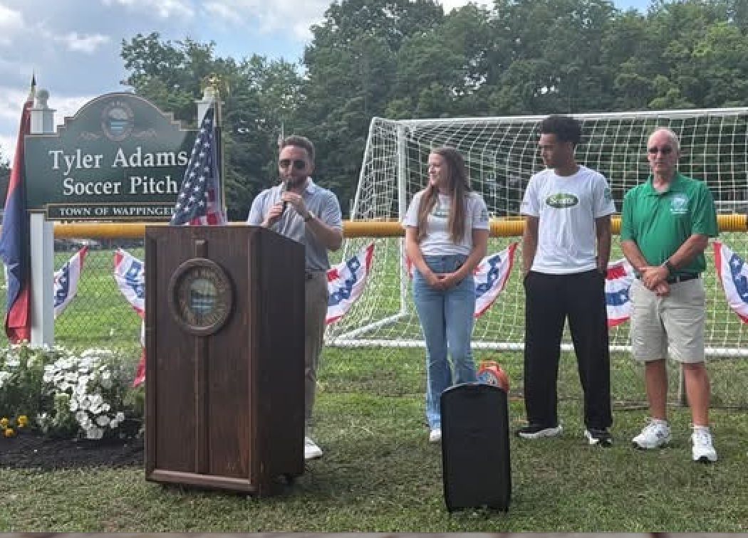 People speaking at a podium in Tyler Adams Soccer Pitch park, with soccer goal and sign in background