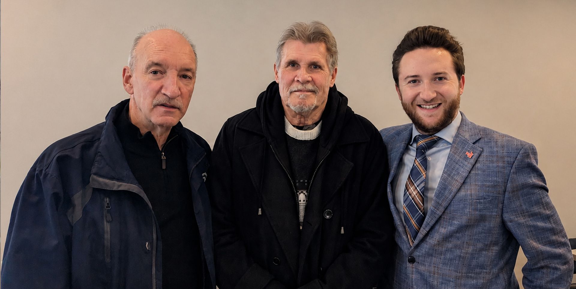 Three men posing indoors, one in a gray suit and two in dark jackets, against a plain wall.
