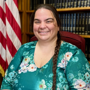 Smiling woman seated in an office, wearing a teal floral blouse with bookshelves and an American flag behind her