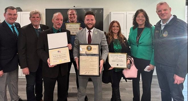 Group of seven people posing indoors with framed certificates and awards, smiling in a formal recognition ceremony