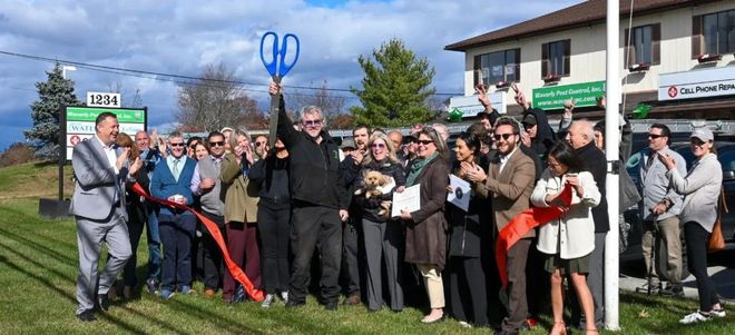 Group of people posing outdoors holding large scissors at a ribbon-cutting ceremony in front of a building.