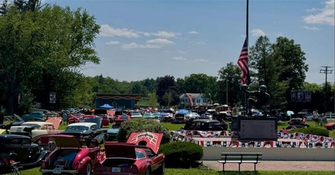 Classic car show with colorful vintage cars, a large American flag, and spectators on a sunny day.