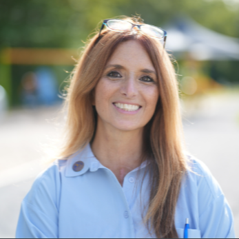 Smiling person in a light blue shirt outdoors with blurred greenery in the background