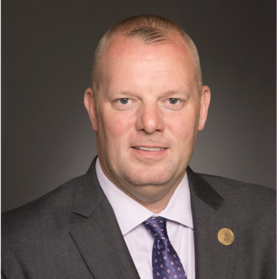 Studio headshot of a man in a gray suit and purple tie against a dark background