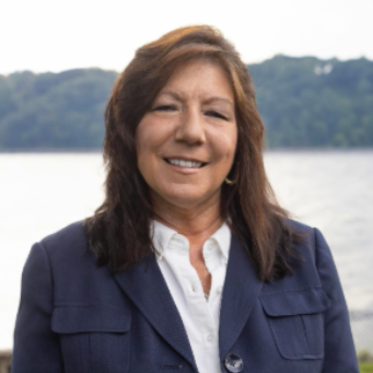 Smiling person in a navy jacket standing outdoors by a lake with trees in the background