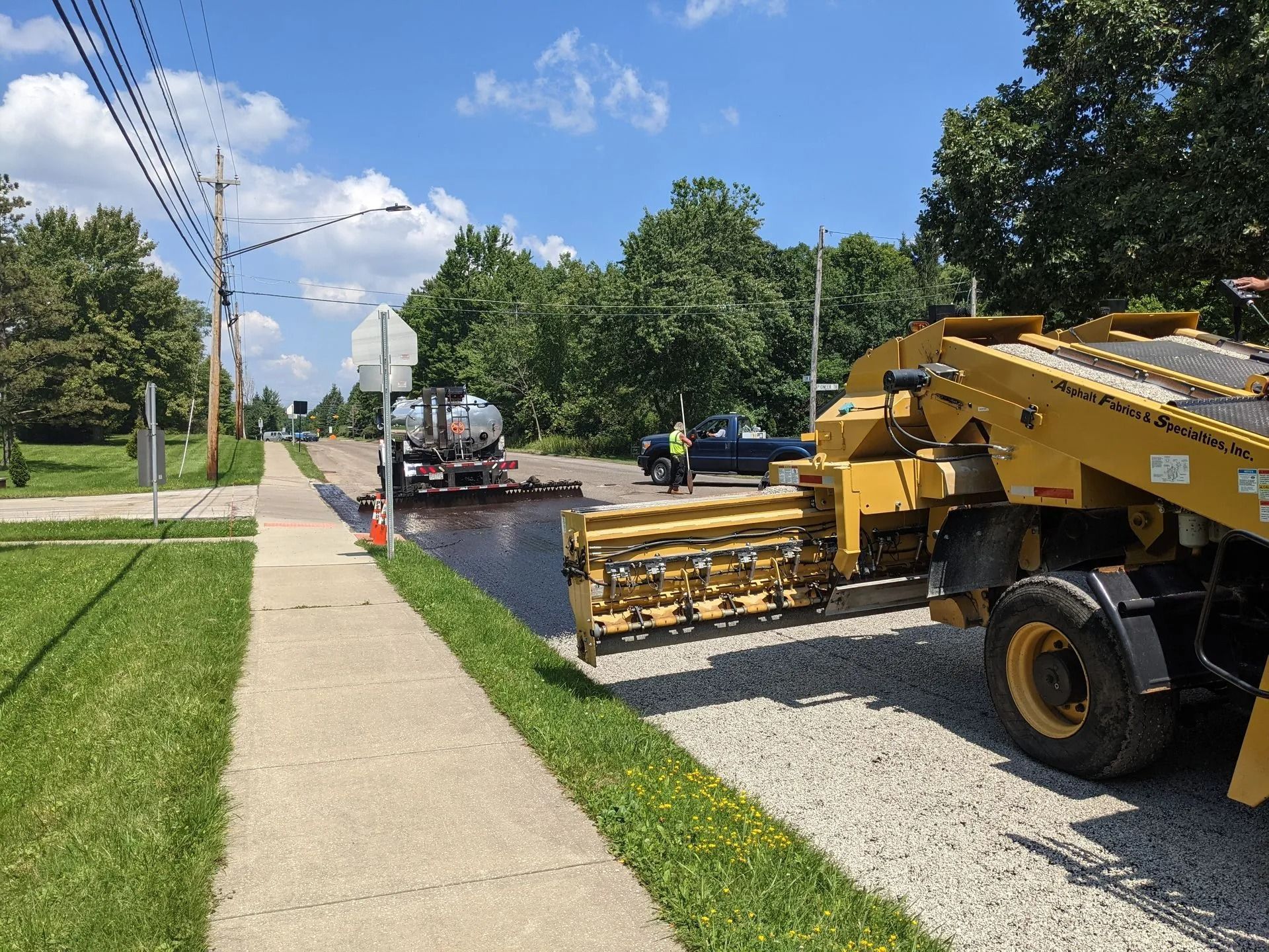 Road resurfacing in progress; asphalt paver lays new blacktop on a residential street; truck and worker present.