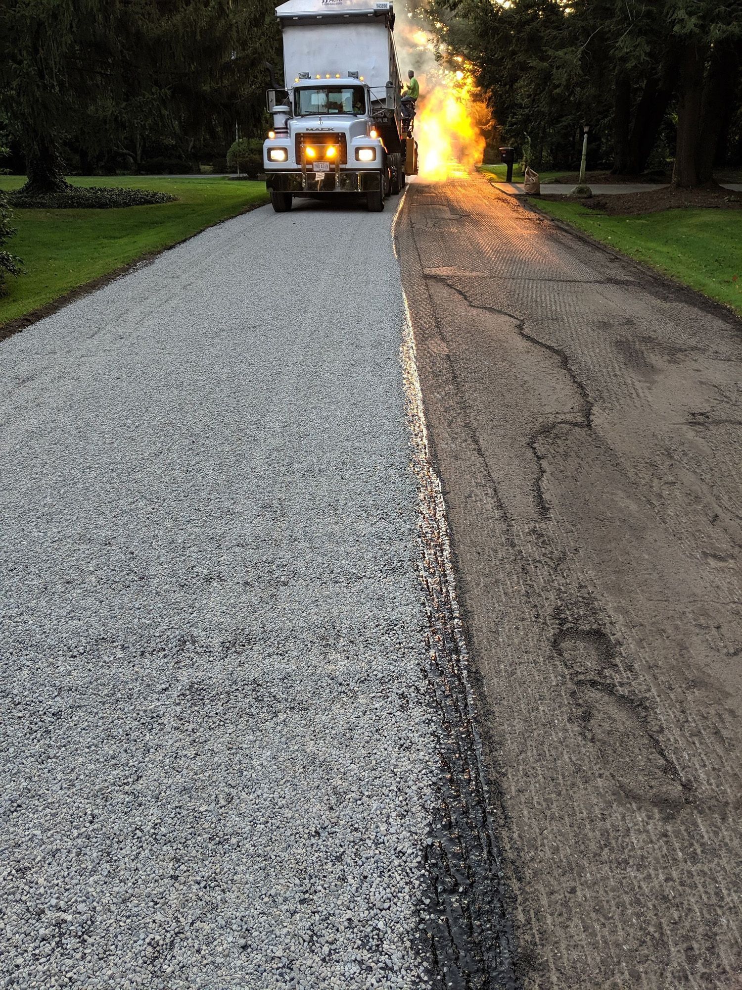 Truck laying gravel on asphalt road, half-completed. The sun shines brightly in the background.