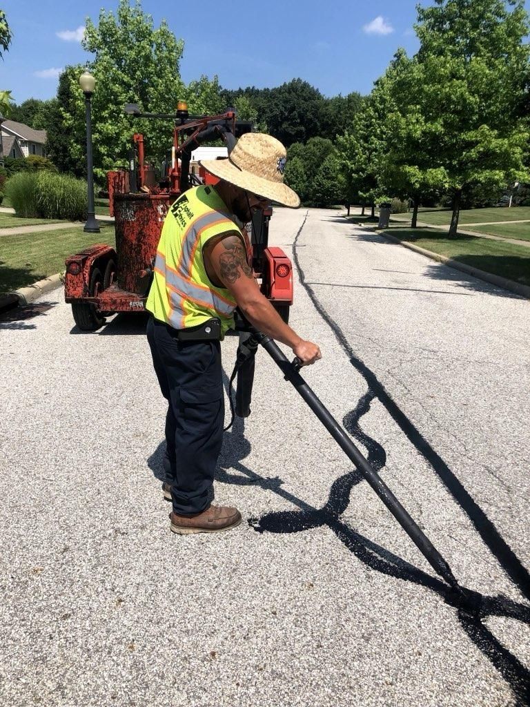 Road worker applying black sealant to asphalt street. He wears a reflective vest and straw hat, with a machine in the background.