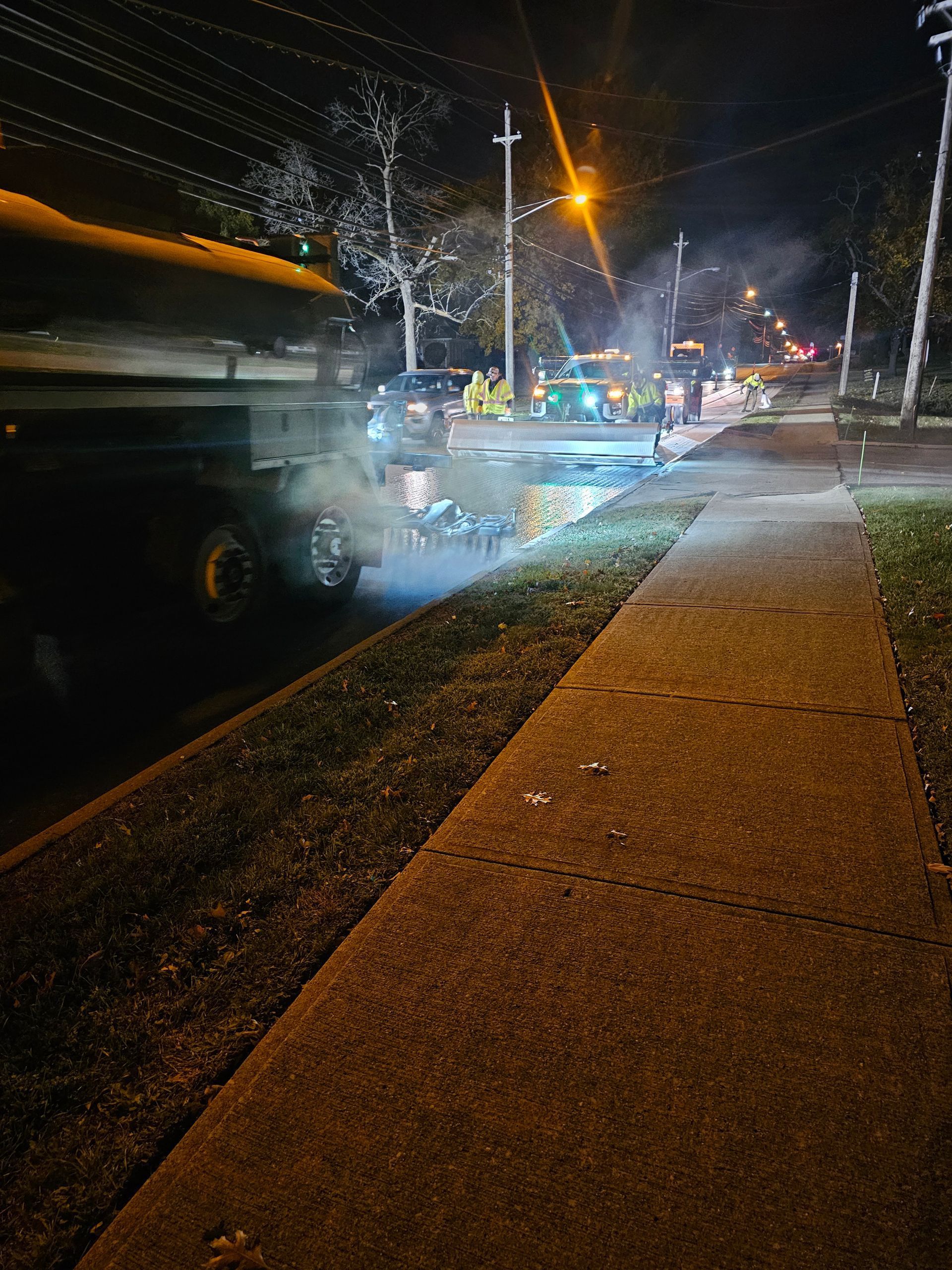 A semi-truck parked at night, smoke coming from the tires on a sidewalk. Streetlights illuminate the area.