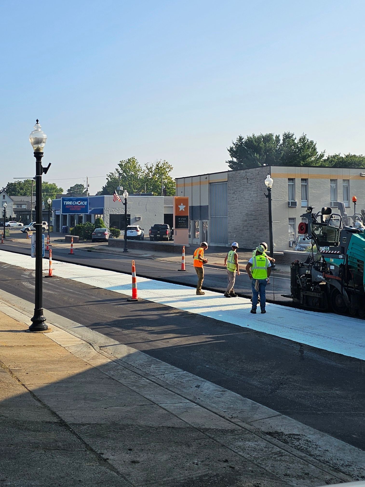 Road construction: Workers in safety vests supervise asphalt paving on a city street; buildings in background.
