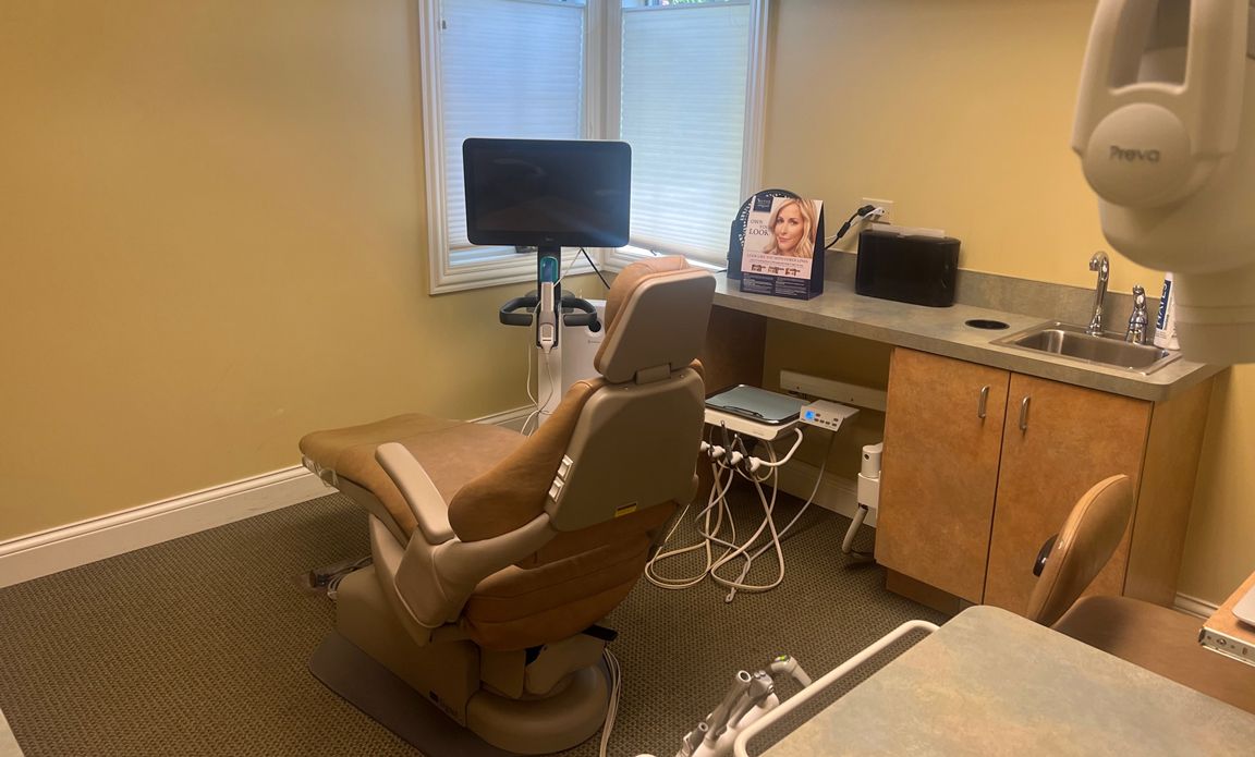 A dental office with a dental chair, desk, sink, and monitor.