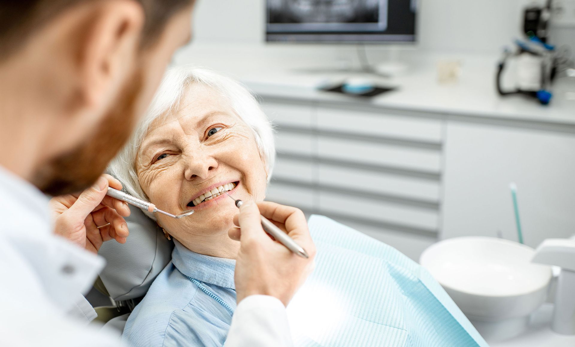 An elderly woman is sitting in a dental chair while a dentist examines her teeth.