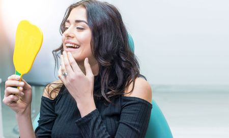 A woman is sitting in a dental chair looking at her teeth in a mirror.