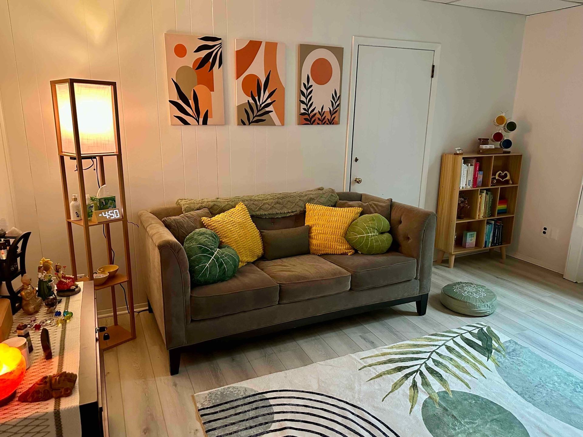 Cozy living room with gray sofa, green pillows, floor lamp, wall art, and a leafy rug by a bookshelf