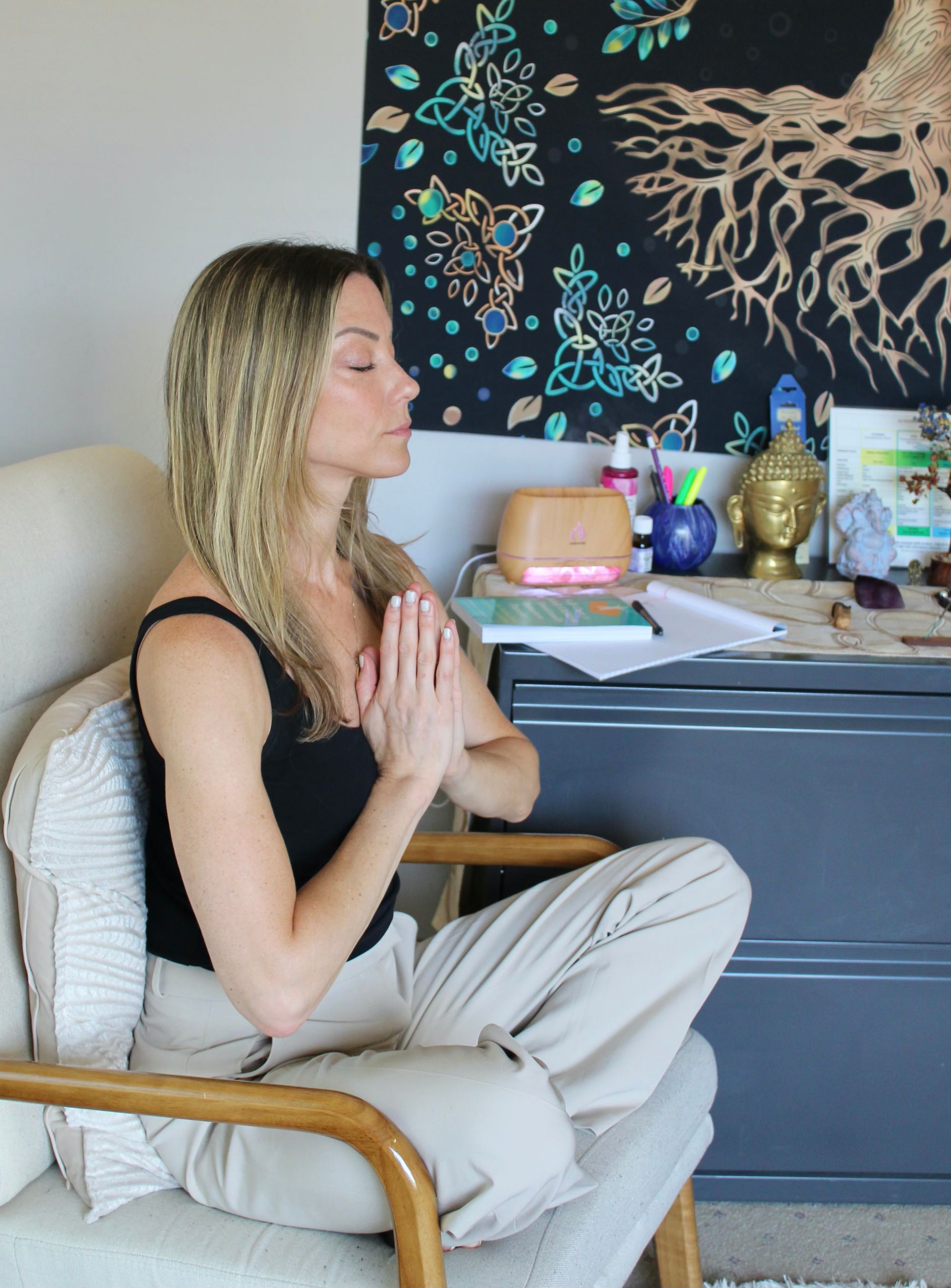 Woman meditating with hands together, sitting in a chair, eyes closed. Neutral setting, artwork and desk in the background.