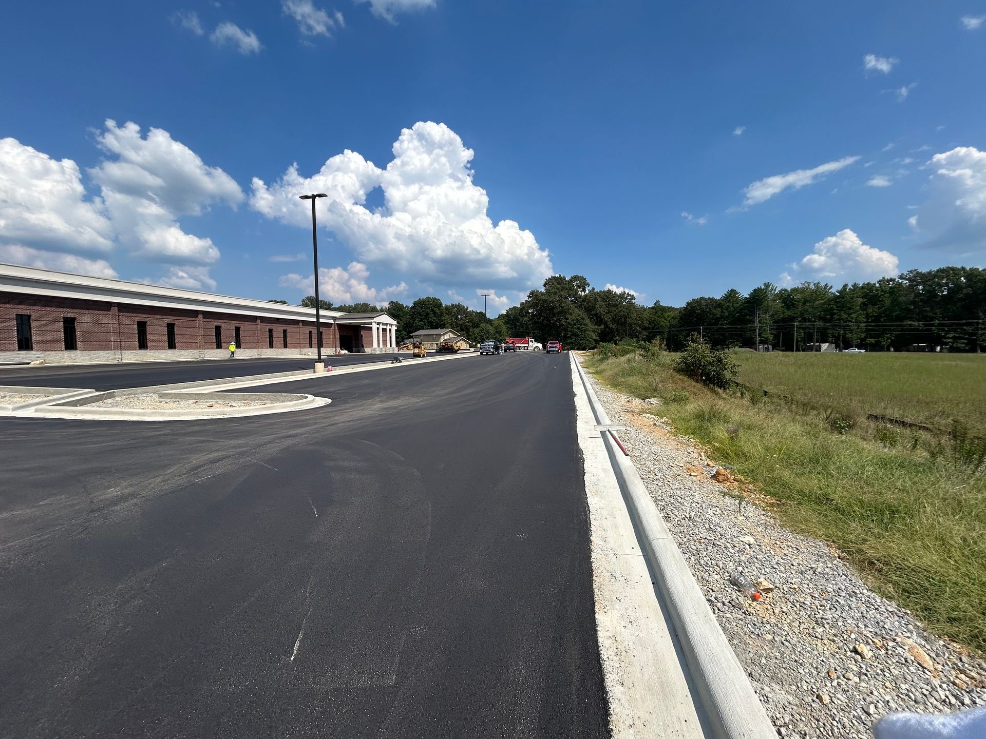 Newly paved road beside a long brick building and grassy field under a blue sky with clouds