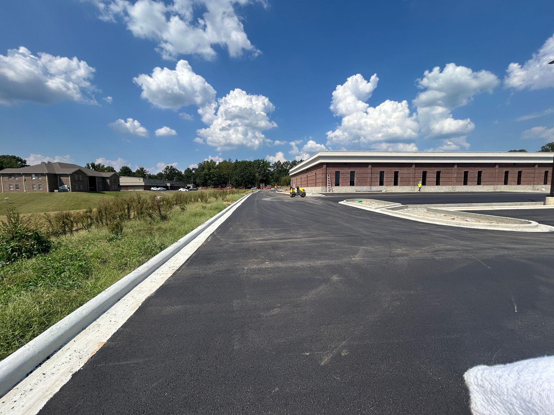 Paved road beside a brick building and grassy field under a bright blue sky with clouds