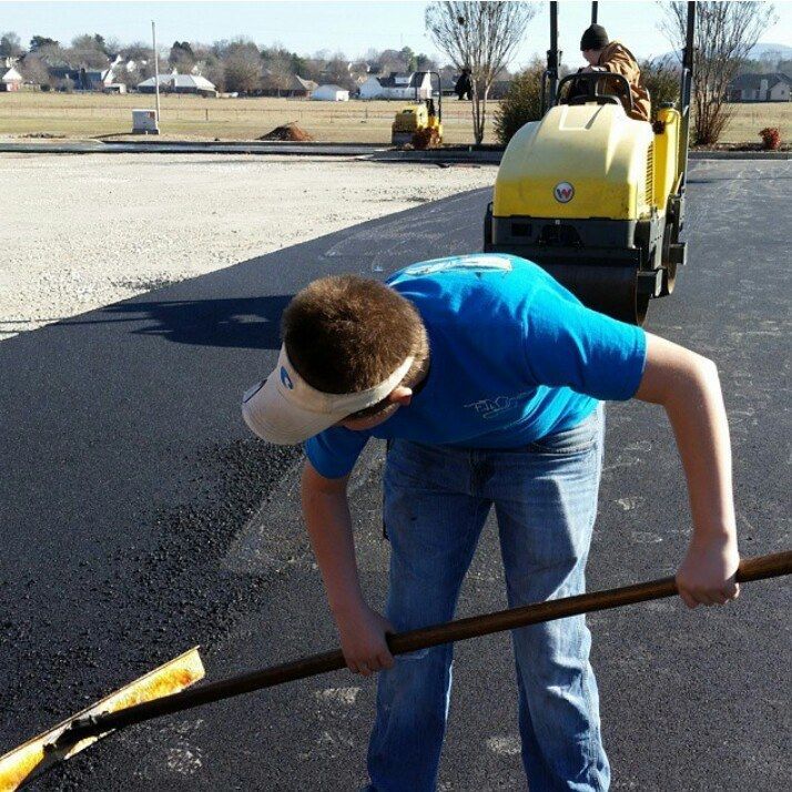 Worker shoveling fresh blacktop beside a yellow road roller on a paved lot