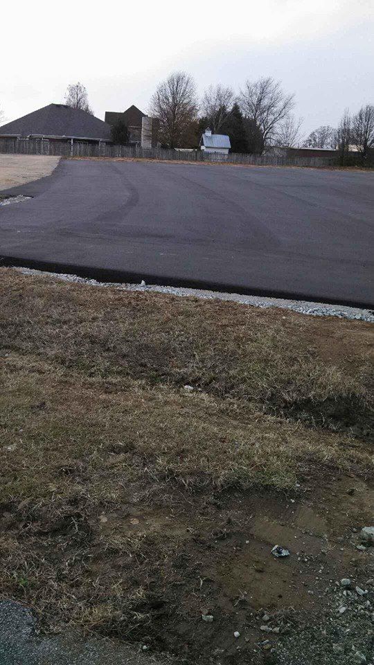 Empty paved lot beside a road with a small building and trees under an overcast sky