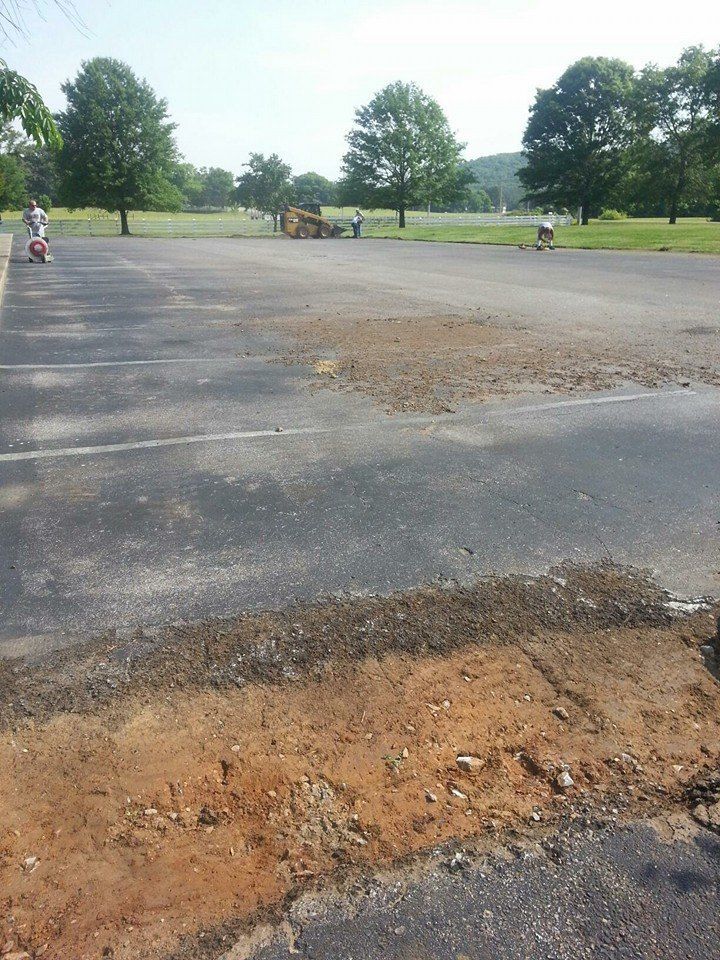 Cracked asphalt parking lot with large potholes and scattered gravel in a park area.