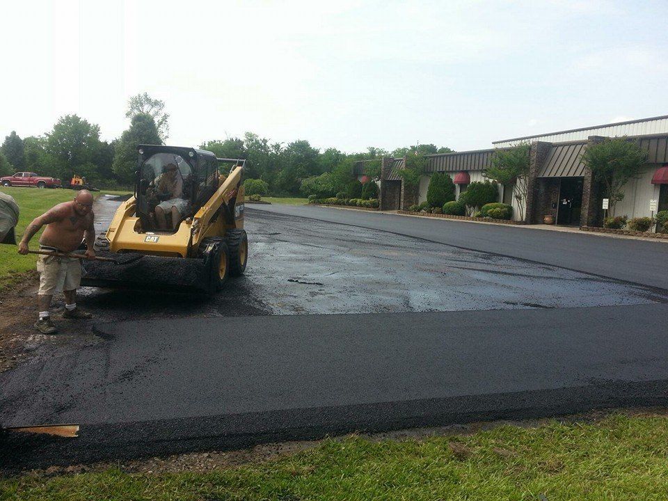 Worker operating a skid steer to spread fresh black asphalt in a parking lot