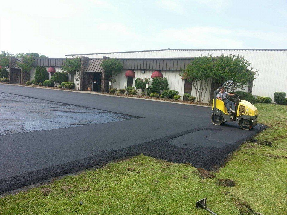 Yellow paving machine laying fresh black asphalt beside a building and grass verge