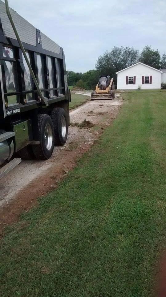 Dump truck unloading gravel for a driveway beside a white house in a grassy yard