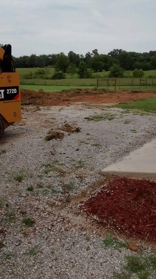 Gravel lot beside a field with trees, partly covered by reddish dirt piles and a yellow machine on the left.