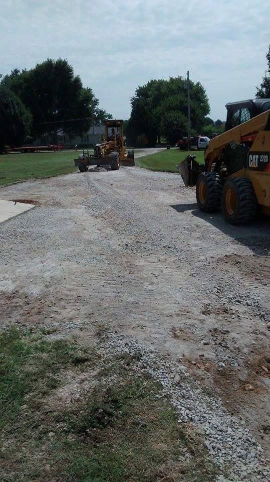 Construction equipment grading a gravel road in a park-like area with trees.