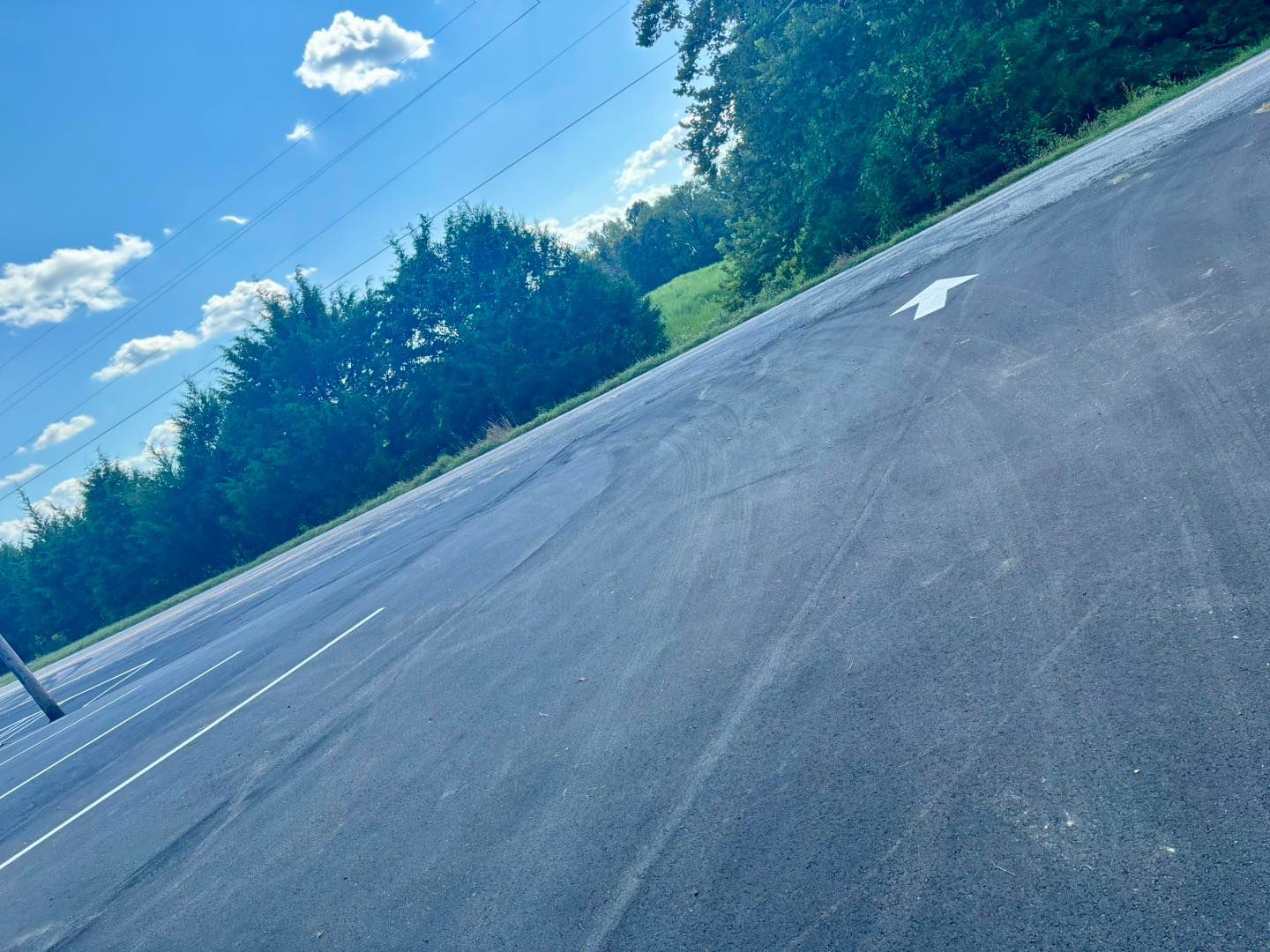 Empty paved parking lot beside trees under a blue sky with clouds