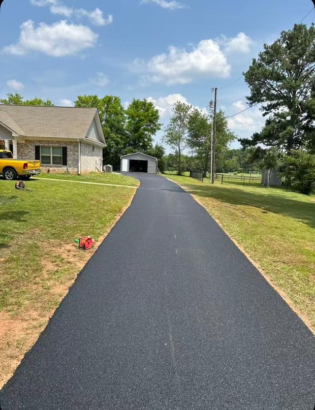 Fresh black asphalt driveway leading to a house and garage on a sunny suburban lot, with a yellow truck on the left.