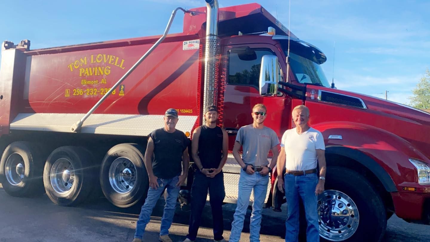 Three people standing beside a red dump truck with yellow lettering on a sunny day.