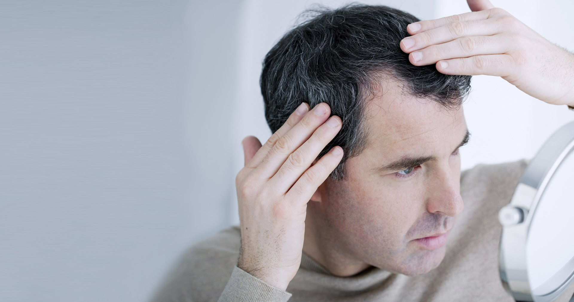 Man examining hairline in a mirror, grey hair visible, touching his head with both hands.